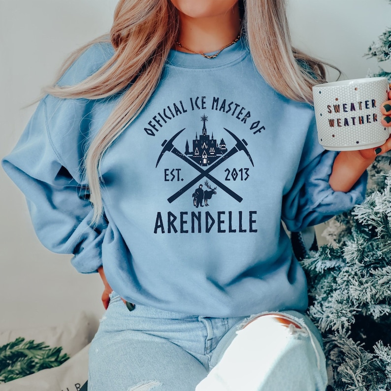 May include: A light blue sweatshirt with a graphic design featuring a castle, crossed pickaxes, and the text "Official Ice Master of Arendelle Est. 2013".