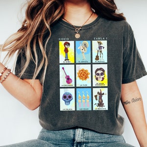 May include: A dark gray t-shirt with a colorful graphic of a "Coco" themed Loteria card game. The graphic features characters and objects from the movie, including Miguel, the guitar, the flower, and the alebrije. The text "COCO" and "TABLA I" are printed at the top of the graphic.