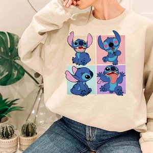 May include: A beige sweatshirt featuring a colorful graphic of the character Stitch from the animated film Lilo & Stitch. The graphic shows Stitch in four different poses, each in a separate square. The sweatshirt is made of a soft material.