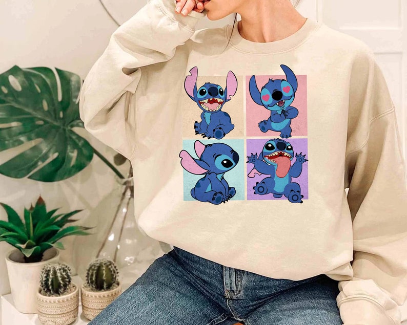 May include: A beige sweatshirt featuring a colorful graphic of the character Stitch from the animated film Lilo & Stitch. The graphic shows Stitch in four different poses, each in a separate square. The sweatshirt is made of a soft material.