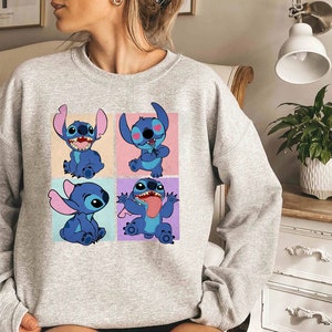 May include: A light gray sweatshirt featuring a four-panel graphic of the character Stitch from Lilo & Stitch. Each panel shows Stitch in a different pose against a pastel background. The sweatshirt is made of a soft material.
