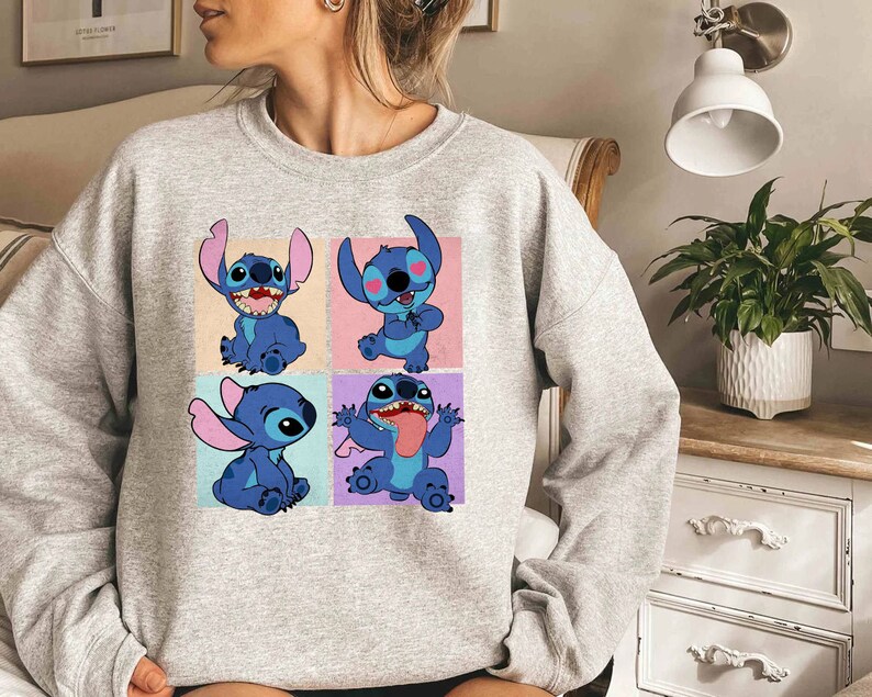 May include: A light gray sweatshirt featuring a four-panel graphic of the character Stitch from Lilo & Stitch. Each panel shows Stitch in a different pose against a pastel background. The sweatshirt is made of a soft material.