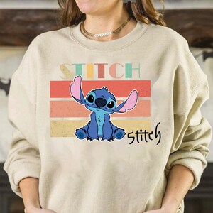 May include: A beige sweatshirt with a retro-style graphic of Stitch from Lilo & Stitch. The graphic features Stitch sitting with his arms crossed and the text "Stitch" in a vintage font.