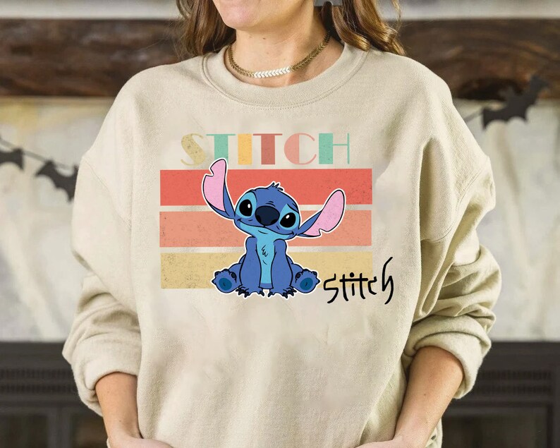 May include: A beige sweatshirt with a retro-style graphic of Stitch from Lilo & Stitch. The graphic features Stitch sitting with his arms crossed and the text "Stitch" in a vintage font.