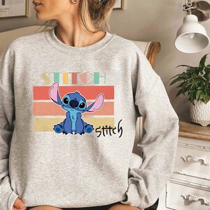 May include: Gray sweatshirt with a blue Stitch character from Lilo & Stitch. The character is sitting on a colorful striped background with the word "Stitch" written below.