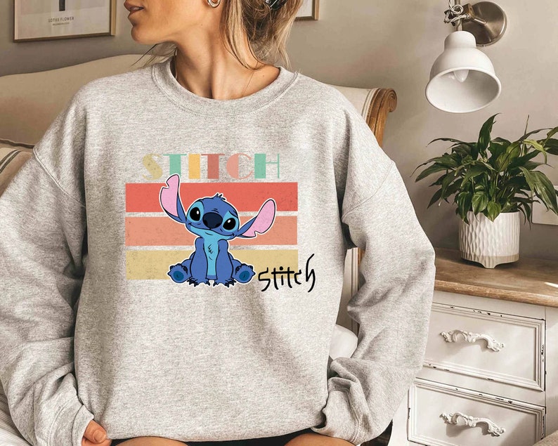 May include: Gray sweatshirt with a blue Stitch character from Lilo & Stitch. The character is sitting on a colorful striped background with the word "Stitch" written below.