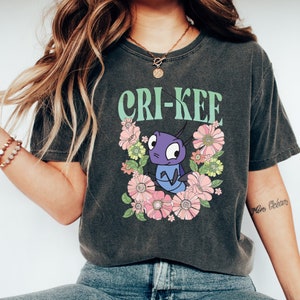 May include: A dark gray t-shirt with a graphic print of a cartoon cricket with the text "CRI-KEE" in green. The cricket is surrounded by pink and yellow flowers.