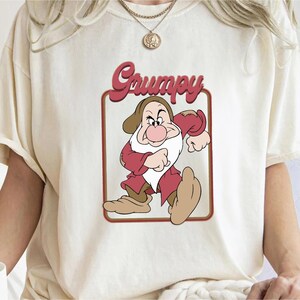 May include: A white t-shirt featuring a red and brown graphic of Grumpy from Snow White. The graphic depicts Grumpy with a grumpy expression and the word "Grumpy" in a retro font.