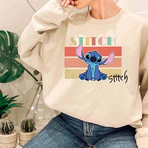 May include: A beige sweatshirt with a retro-style graphic of Stitch from Lilo & Stitch. The graphic features Stitch in blue with a pink ear and a red background. The text "Stitch" is written in a colorful font above and below the graphic.
