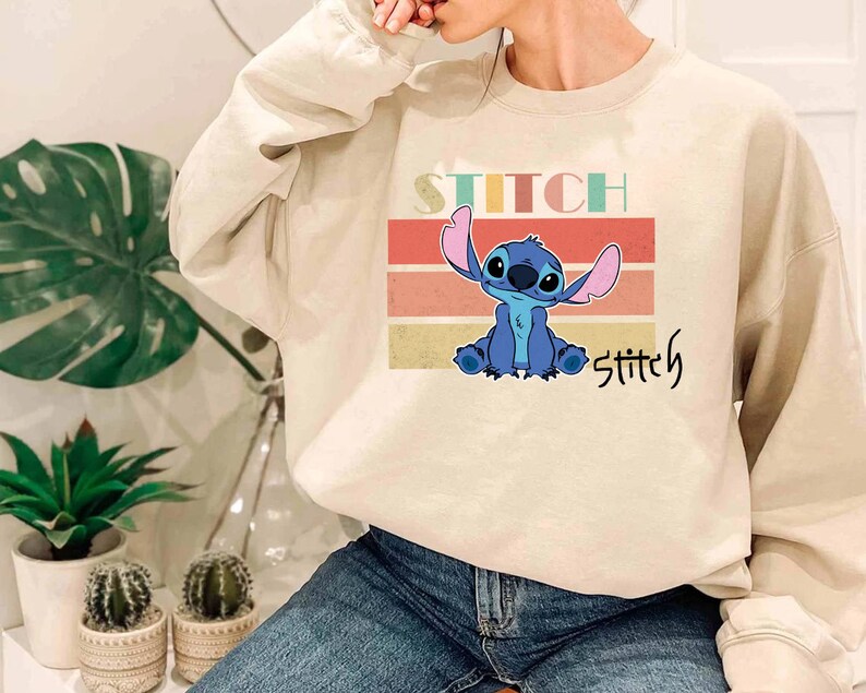 May include: A beige sweatshirt with a retro-style graphic of Stitch from Lilo & Stitch. The graphic features Stitch in blue with a pink ear and a red background. The text "Stitch" is written in a colorful font above and below the graphic.
