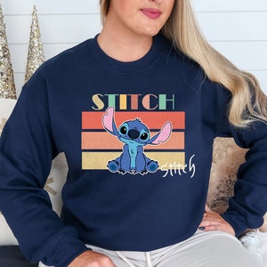May include: A navy blue sweatshirt with a retro-style graphic of Stitch from Lilo & Stitch. The graphic features Stitch sitting with his arms raised and the text "Stitch" above him and "Stitchly" below him.