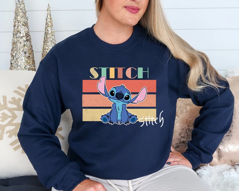 May include: A navy blue sweatshirt with a retro-style graphic of Stitch from Lilo & Stitch. The graphic features Stitch sitting with his arms raised and the text "Stitch" above him and "Stitchly" below him.