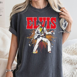 May include: A dark gray t-shirt with a vintage-style graphic of Elvis Presley performing on stage. The graphic features Elvis in a white jumpsuit with a black belt and a microphone in his hand. The text "ELVIS" is printed in red above the graphic.