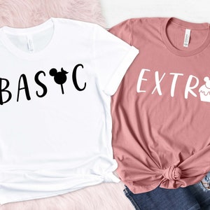 Peut inclure: Deux t-shirts, un blanc et un rose poudré, avec du texte noir et blanc. Le t-shirt blanc dit "BASIC" avec une icône de tête de Mickey Mouse. Le t-shirt rose poudré dit "EXTRA" avec une icône de cupcake. Les deux t-shirts sont à manches courtes.