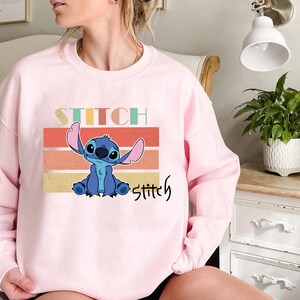 May include: Pink sweatshirt with a blue Stitch character from Lilo & Stitch. The text "Stitch" is printed in a retro font on the sweatshirt.