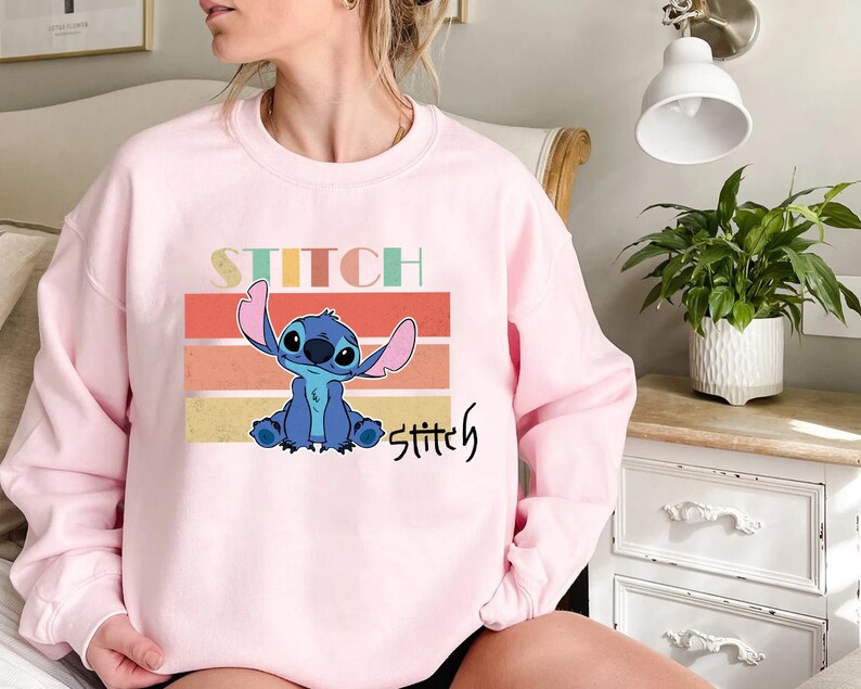 May include: Pink sweatshirt with a blue Stitch character from Lilo & Stitch. The text "Stitch" is printed in a retro font on the sweatshirt.