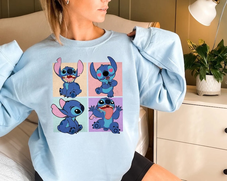 May include: A light blue sweatshirt featuring a four-panel design of the character Stitch from the animated film Lilo & Stitch. Each panel showcases a different pose of Stitch, with varying expressions and gestures, all in shades of blue.
