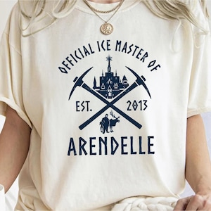 May include: A white t-shirt with a blue graphic design. The graphic features a castle, crossed pickaxes, and the text "Official Ice Master of Arendelle Est. 2013".