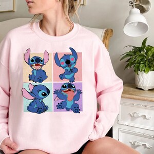 May include: A light pink sweatshirt featuring a four-panel graphic of the character Stitch from Lilo & Stitch. Each panel shows Stitch in a different pose, with a blue color scheme and various background colors.