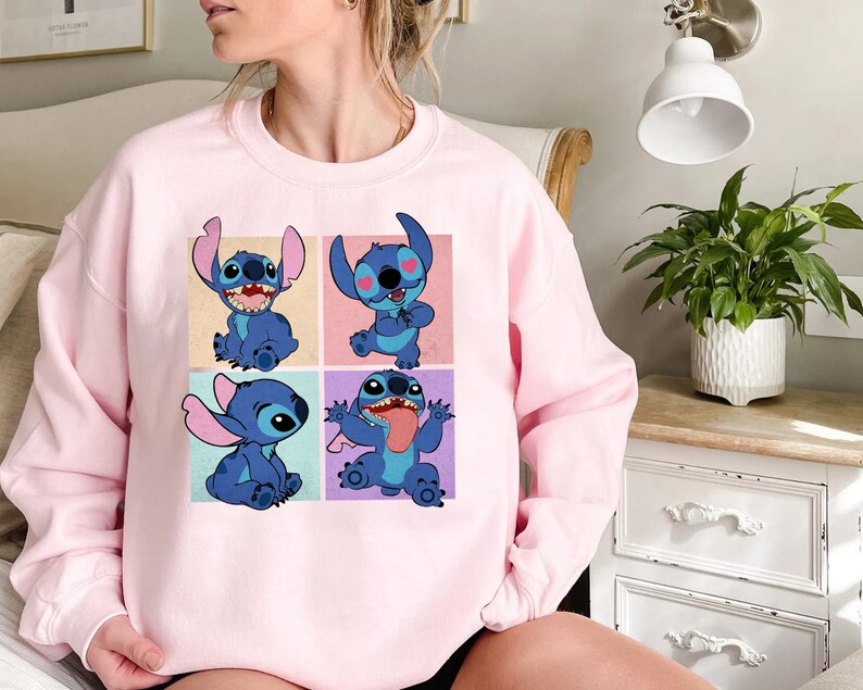 May include: A light pink sweatshirt featuring a four-panel graphic of the character Stitch from Lilo & Stitch. Each panel shows Stitch in a different pose, with a blue color scheme and various background colors.