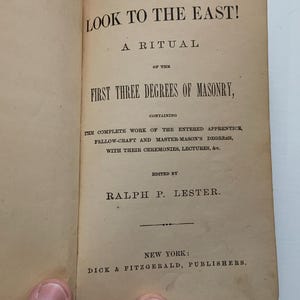 Peut inclure: Un livre ouvert aux pages jaunies et blanc cassé avec du texte imprimé en noir. La page de titre indique : LOOK TO THE EAST! A RITUAL OF THE FIRST THREE DEGREES OF MASONRY, CONTAINING THE COMPLETE WORK OF THE ENTERED APPRENTICE, FELLOW-CRAFT.