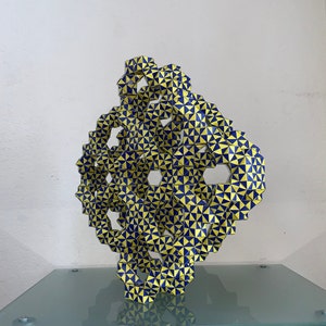 Könnte beinhalten: Eine geometrische Skulptur aus miteinander verbundenen Dreiecksformen. Die Skulptur weist ein sich wiederholendes Muster aus gelben und marineblauen Dreiecken auf. Sie wird auf einer Glasoberfläche ausgestellt.