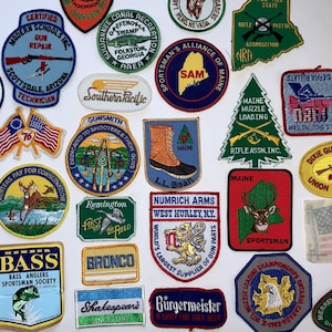 Op de afbeelding: Een verzameling geborduurde patches met verschillende ontwerpen en tekst. Patches bevatten logo's voor wapenreparatie, jagen, sport en bedrijven. Kleuren zijn rood, blauw, groen en geel. Sommige patches tonen vlaggen en dieren in het wild.