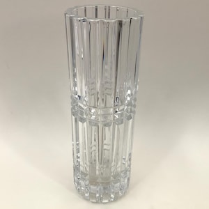 May include: A clear, tall glass vase with vertical, ridged detailing. The vase has a rectangular shape with a slightly flared top edge. The glass is transparent, allowing light to pass through. The vase is likely designed for displaying flowers.
