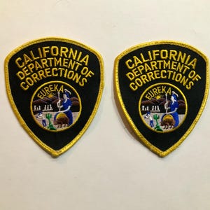 Peut inclure: Deux écussons brodés en forme de bouclier noir avec une bordure dorée. Les écussons portent l'inscription "CALIFORNIA DEPARTMENT OF CORRECTIONS" en lettres dorées. Chaque écusson comporte un emblème circulaire avec une femme, un ours et le mot "EUREKA".