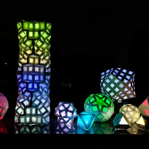 May include: A collection of illuminated geometric paper lanterns in various shapes and sizes. The lanterns are lit from within, displaying a spectrum of colors including green, blue, and red. The tallest lantern is a tall, rectangular tower.