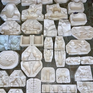 Puede incluir: Una colección de moldes de yeso blanco de varias formas y tamaños, incluyendo figuras, relieves y tazas. Los moldes representan escenas y personajes detallados, probablemente para crear artículos decorativos o esculturas. La disposición sugiere un taller o un entorno artesanal.
