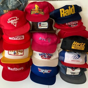 Pode incluir: Uma coleção de bonés de caminhoneiro vintage em várias cores, incluindo vermelho, azul e preto. Os bonés apresentam logotipos e textos de marcas como Toyota, Marlboro e Hertz. Alguns bonés têm costas de malha.