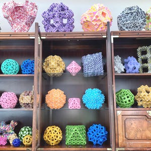 May include: A wooden cabinet with glass doors displays a collection of colorful origami geometric shapes. The shapes are arranged on shelves and include spheres, cubes, and other geometric forms.
