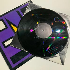 Może przedstawiać: Czarna płyta winylowa z holograficznym wzorem, na okładce albumu. Okładka albumu jest fioletowa z białymi i żółtymi geometrycznymi kształtami. Album nosi tytuł "True Colours" zespołu Split Enz.