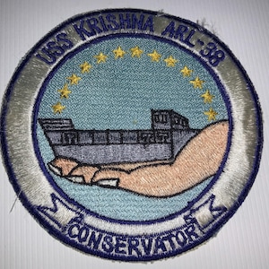 May include: Embroidered patch featuring the text "USS KRISHNA ARL-38" in a circular design. The patch includes a ship illustration held by a hand, with the word "CONSERVATOR" at the bottom. The design is primarily blue, white, and gold.