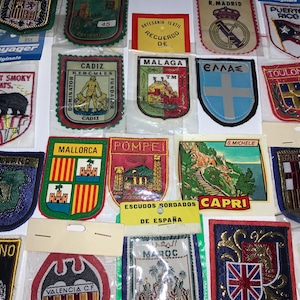 May include: A collection of embroidered patches featuring various locations and symbols. Patches include "Great Smoky Mts.", "Cadiz", "Malaga", "Ireland", "Mallorca", "Pompei", "Capri", and "Puerto Rico". The patches are in various shapes and colors.
