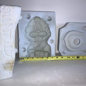 May include: Three light-colored molds, including one with the word "ATLANTIC" embossed on the side. The central mold features a lion design. A yellow measuring tape is placed in front of the molds, showing measurements in inches.