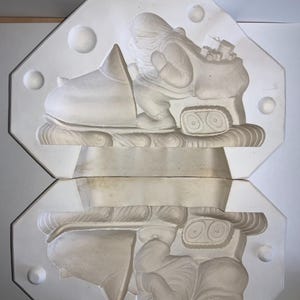 May include: A two-part, off-white plaster mold featuring a Santa Claus figure riding a snowmobile. The Santa figure is carrying a sack of gifts. The mold is in an octagonal shape with decorative spheres on the edges. This item is likely for holiday crafts.