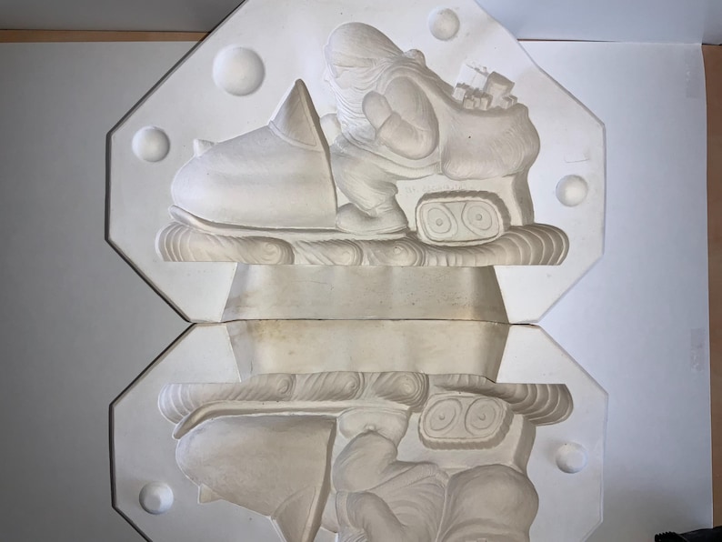 May include: A two-part, off-white plaster mold featuring a Santa Claus figure riding a snowmobile. The Santa figure is carrying a sack of gifts. The mold is in an octagonal shape with decorative spheres on the edges. This item is likely for holiday crafts.