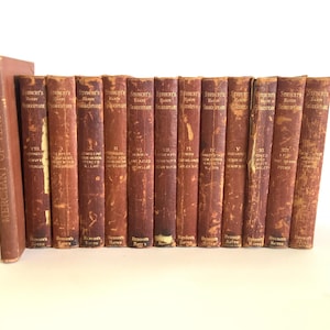 Puede incluir: Una colección de libros antiguos con cubiertas marrones. Las lomos muestran letras doradas, con títulos como "Student's Handbook Shakespeare" y "Merchant of Venice". Los libros están dispuestos en fila, sugiriendo un tema literario.