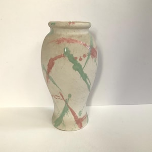 May include: A ceramic vase with a cream-colored base featuring a speckled texture and abstract paint splatters in shades of muted coral pink and sage green. The vase has a rounded body, a narrow neck, and a flared rim with a smooth finish.
