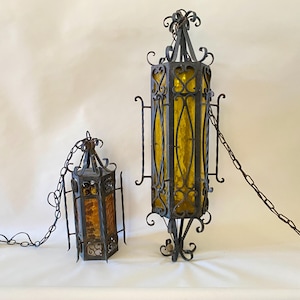 Op de afbeelding: Twee vintage hangende lantaarns van smeedijzer. De lantaarns hebben een donkere afwerking en zijn voorzien van amberkleurige glazen panelen. De grotere lantaarn is rechthoekig, terwijl de kleinere zeshoekig is. Beide zijn opgehangen aan kettingen.