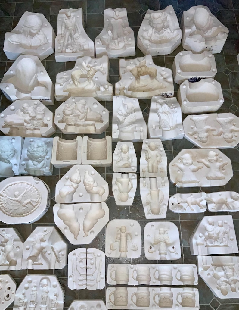 May include: A collection of white plaster molds in various shapes and sizes, including figures, reliefs, and mugs. The molds depict detailed scenes and characters, likely for creating decorative items or sculptures. The arrangement suggests a workshop or craft setting.