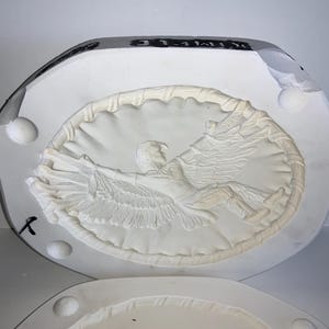 May include: A white plaster mold featuring a detailed relief of an eagle with outstretched wings, encircled by a decorative border. The mold is in two parts, with a partially visible lower section.