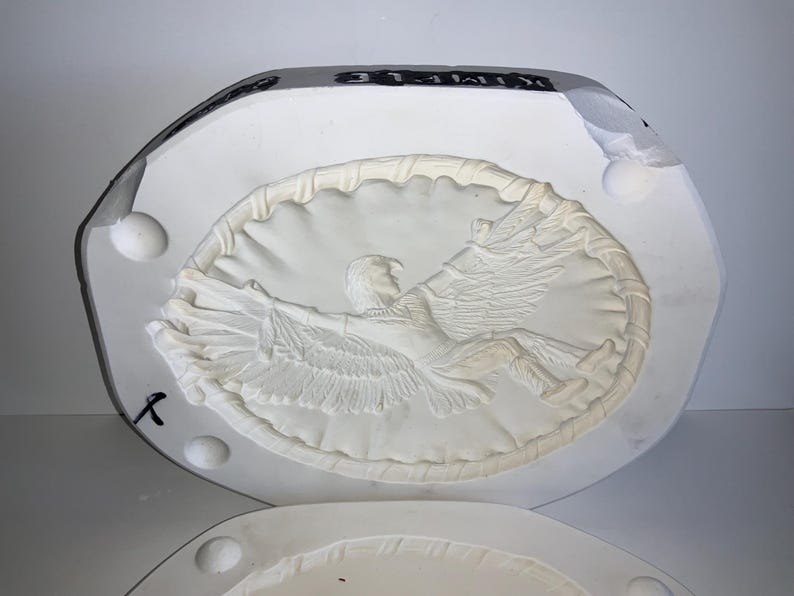 May include: A white plaster mold featuring a detailed relief of an eagle with outstretched wings, encircled by a decorative border. The mold is in two parts, with a partially visible lower section.