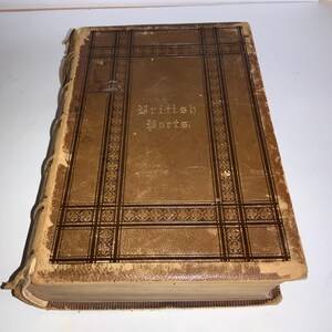 May include: A brown leather-bound book with gold lettering on the cover that reads "British Poets". The cover has a decorative pattern of brown and gold squares and lines.