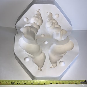 May include: A white ceramic mold with four detailed sections, likely for casting decorative items. The mold is approximately 12 inches long, with a hexagonal shape and rounded edges. The mold is on a white surface.