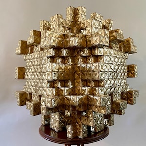 May include: A large, spherical sculpture made from many gold-coloured, cube-shaped elements. The geometric design sits on a dark wooden stand, creating a striking visual contrast. The sculpture's surface reflects light, adding depth and interest.