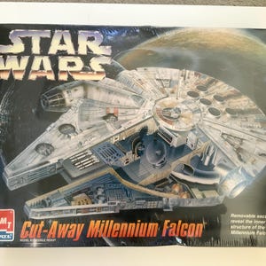 Puede incluir: Una caja de kit de modelo a escala Star Wars Cut-Away Millennium Falcon de AMT Ertl. El empaque presenta una ilustración detallada de la nave espacial con secciones extraíbles que revelan la estructura interna, sobre un fondo espacial oscuro.