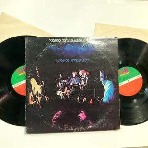 Könnte beinhalten: Ein Vintage-Vinyl-Schallplattenalbum von Crosby, Stills, Nash & Young mit dem Titel "4 Way Street". Das Albumcover zeigt ein dunkles Bild der Band bei der Aufführung. Zwei schwarze Vinylplatten sind sichtbar, mit grünen und roten Etiketten.